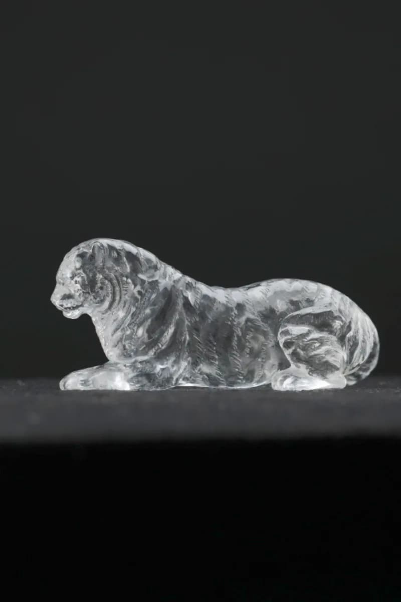 Side profile of a recumbent clear quartz tiger facing left, forepaws extended, against a plain dark grey background; incised fur lines visible along the flank.